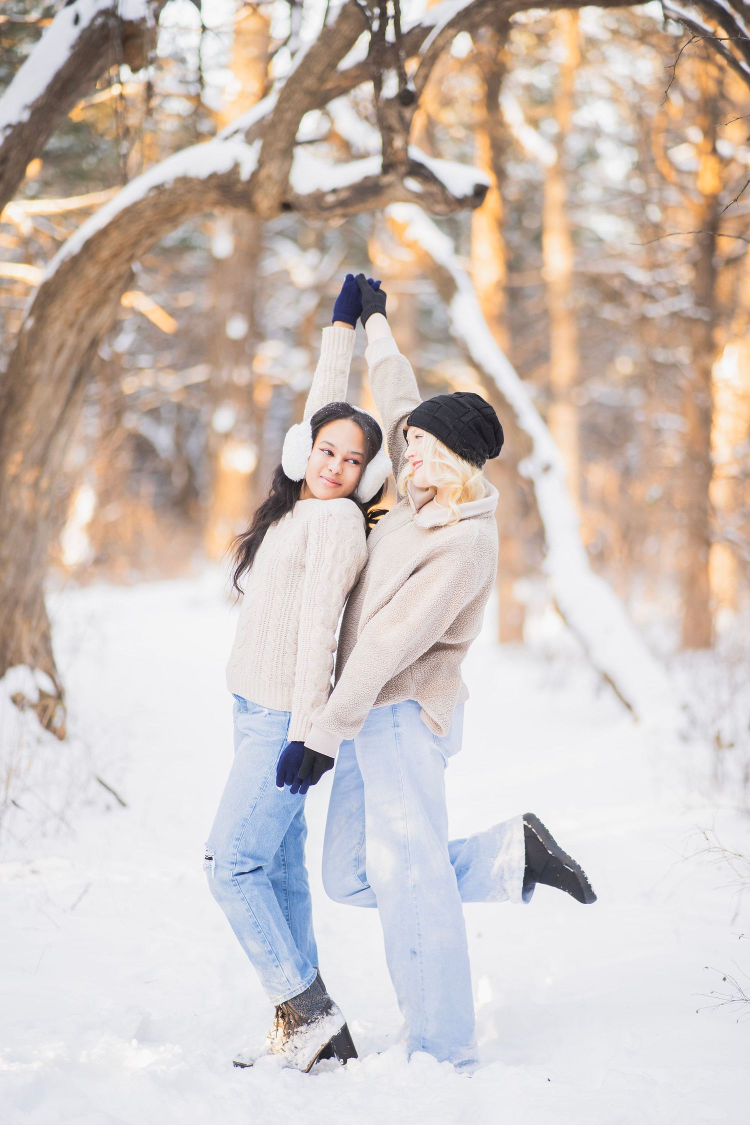 Two young women in beige sweaters laughing and posing together in a snowy winter forest.
