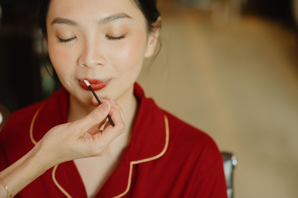 Woman in red pajamas applying red lipstick with eyes closed during makeup routine.
