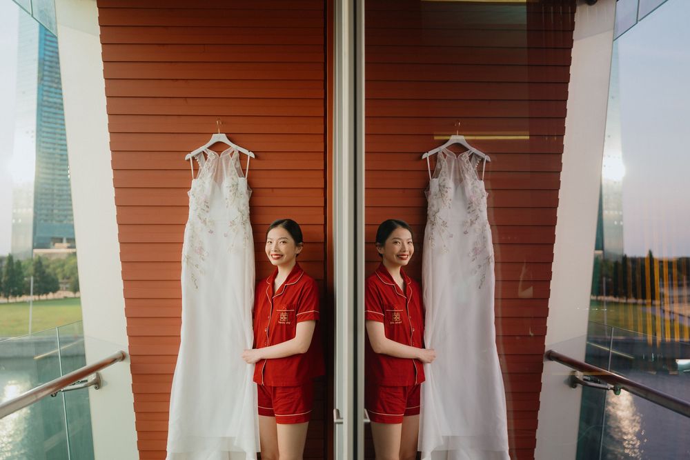 Two bridesmaids in red satin pajamas pose beside hanging white wedding dresses on a glass balcony.