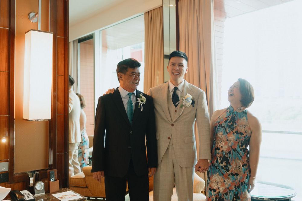 A groom in a beige suit stands smiling with his parents wearing formal attire at an elegant wedding venue.