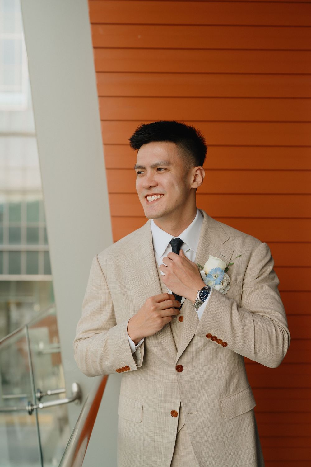 Smiling groom in beige suit adjusting tie with boutonniere, standing against orange wall.