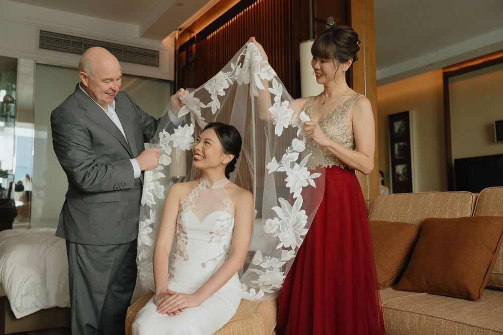 Bride seated while man and woman help arrange floral wedding veil in elegant hotel room.