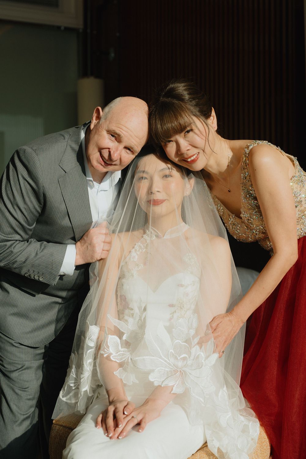 A bride in a white lace gown and veil poses with her smiling parents on her wedding day.