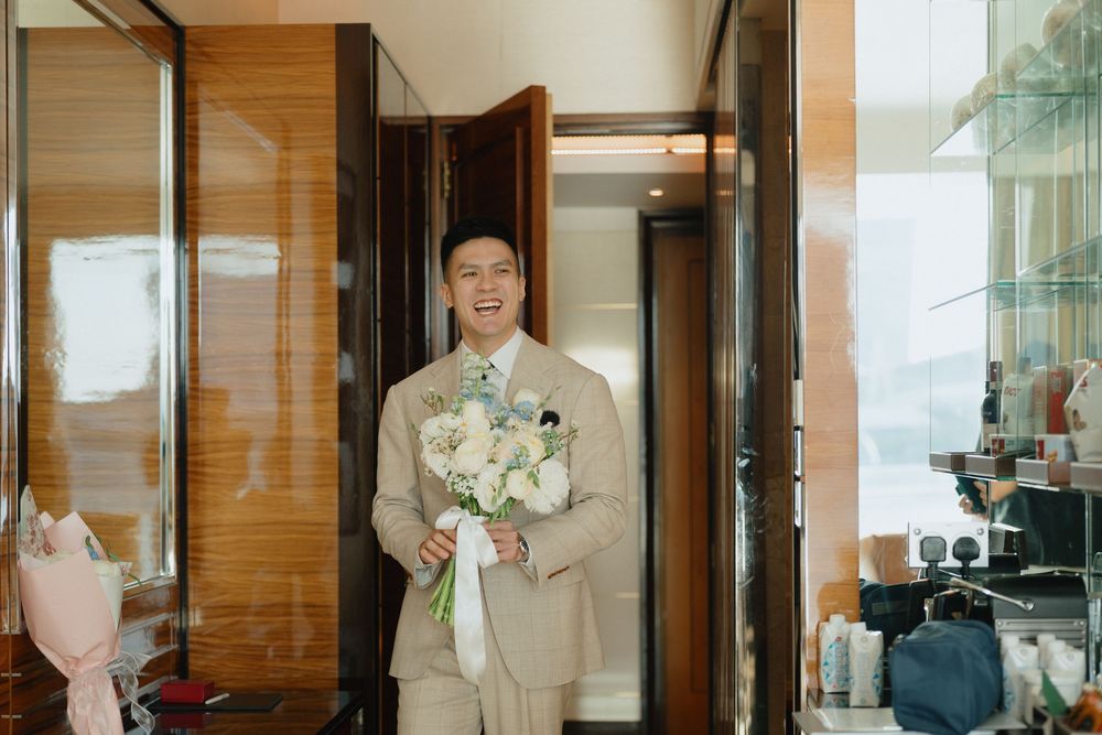 Smiling groom in beige suit holding white floral bouquet standing in elegant hotel room doorway.