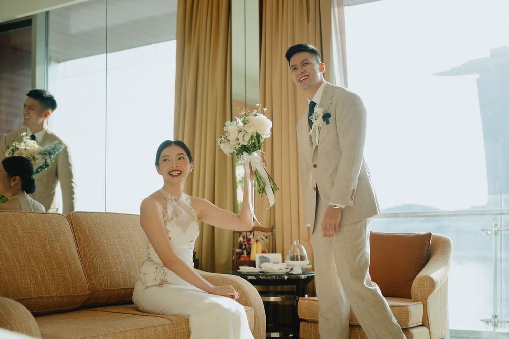 Bride and groom posing in a luxury hotel suite, she holds white flowers while wearing an elegant white dress.
