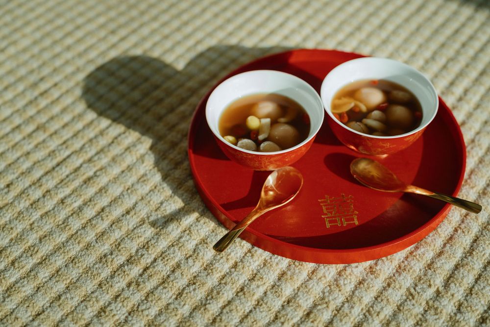 Two bowls of Chinese tang yuan sweet soup on a red tray with gold spoons, served on a textured surface.