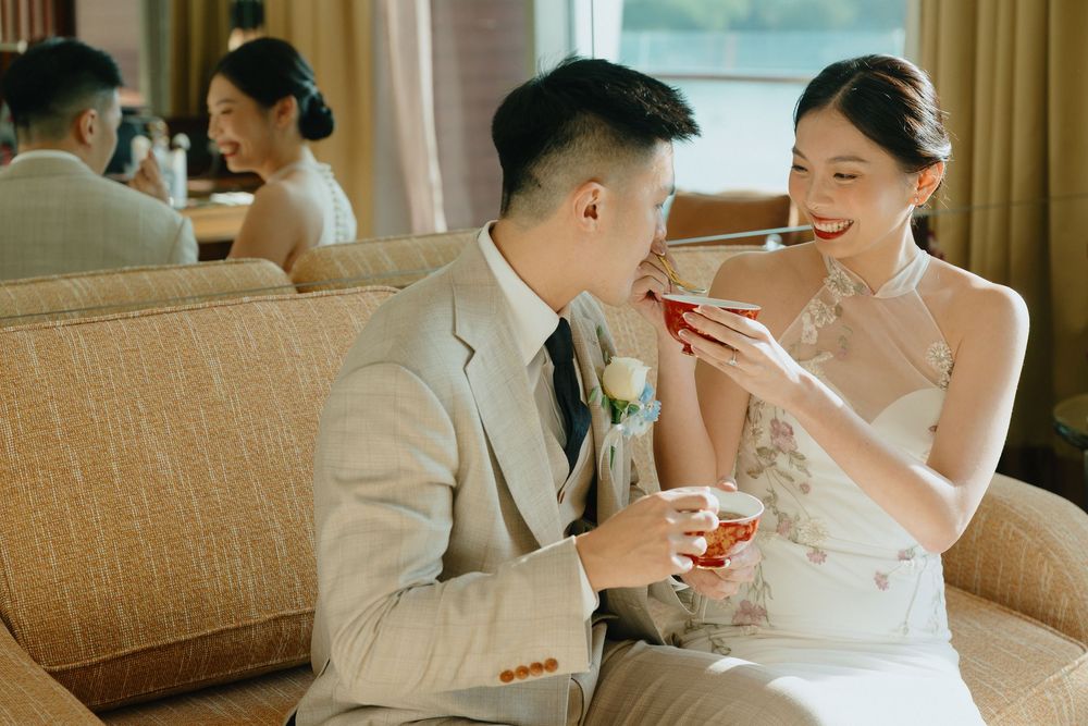Bride in white dress feeds groom in beige suit during wedding reception on elegant sofa.
