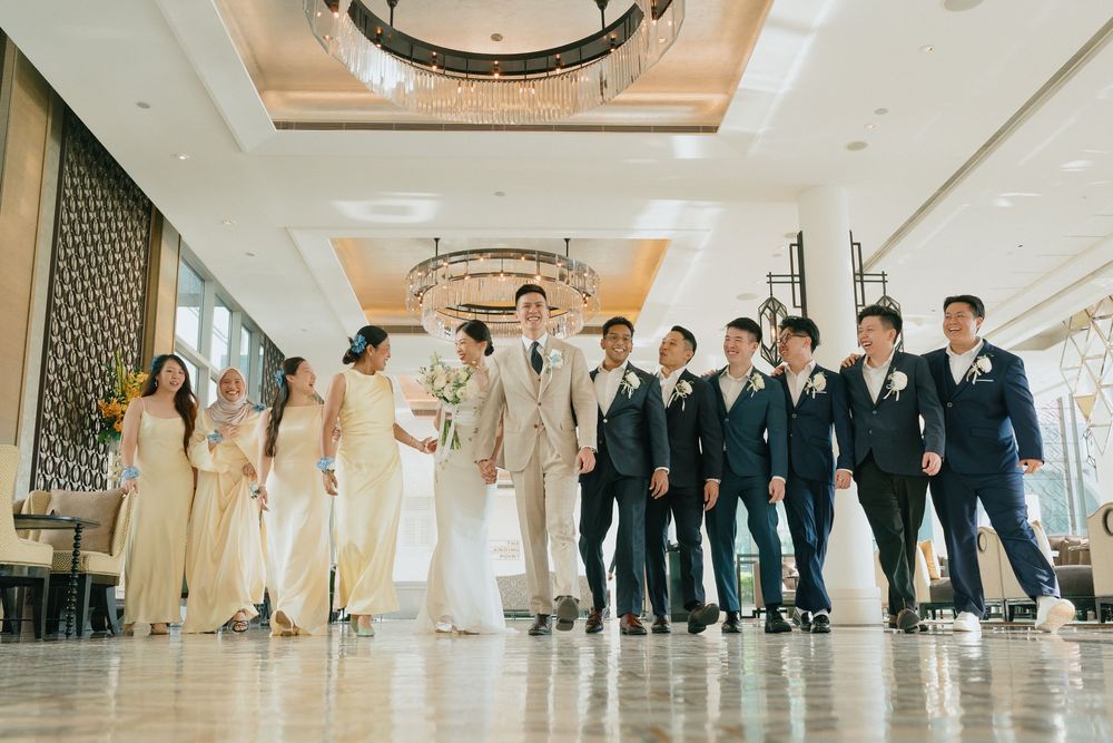 Wedding party poses in elegant hotel lobby, bridesmaids in champagne gowns and groomsmen in navy suits flanking the bride and groom.