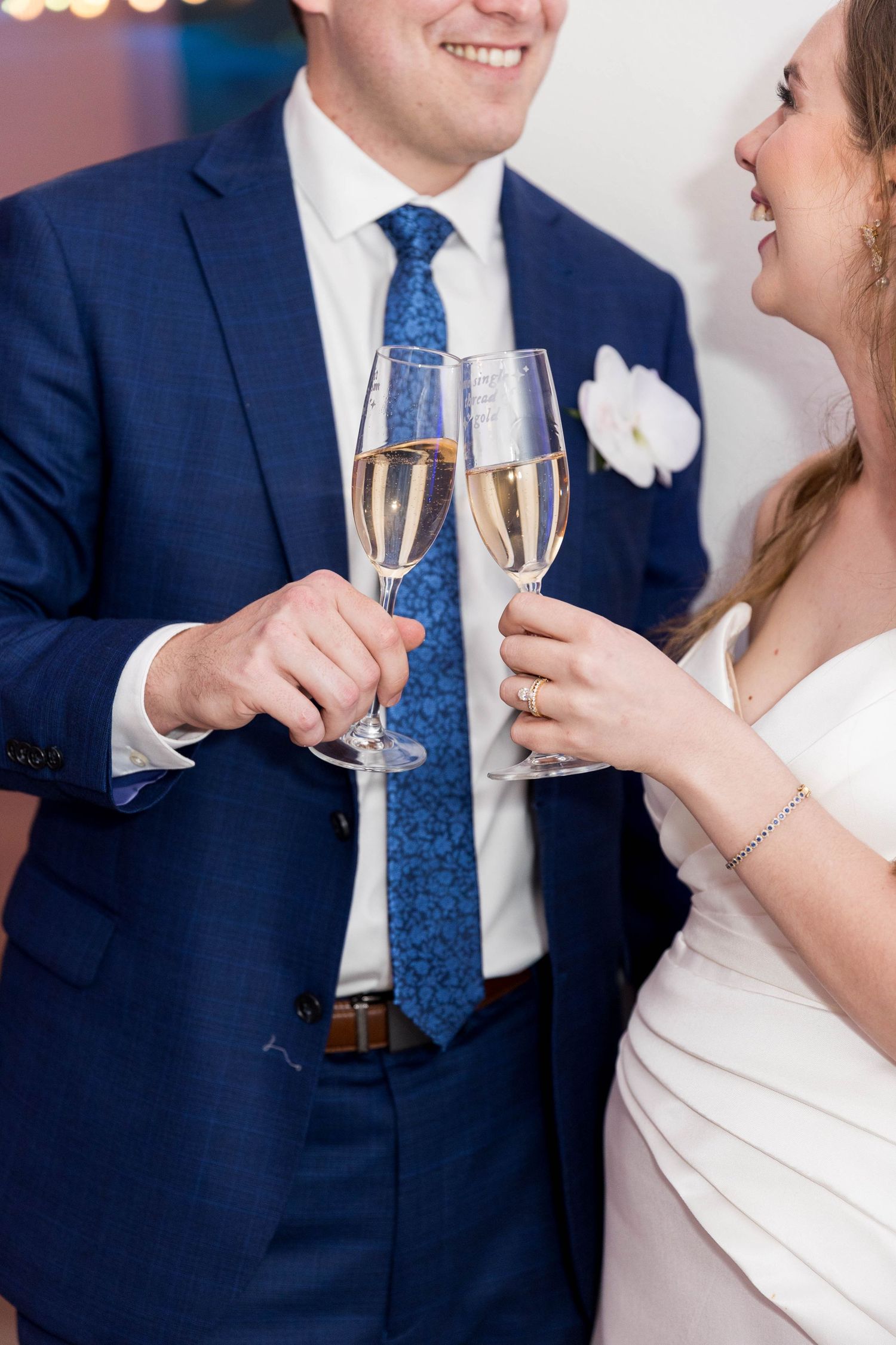 Bride and groom toasting with champagne glasses at wedding reception, man in blue suit with floral boutonniere.