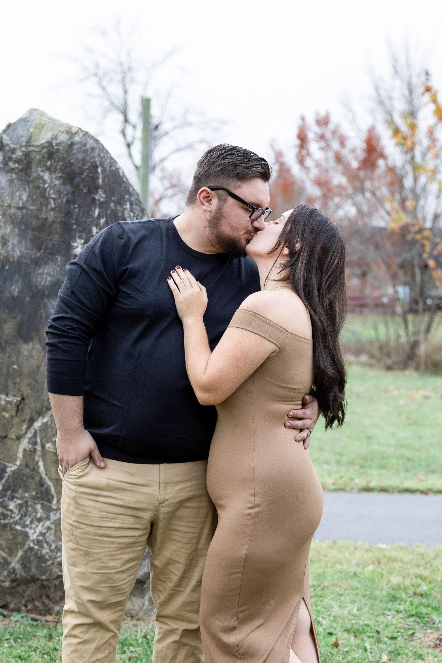 A couple shares a romantic kiss outdoors in a park during autumn, man in black top and woman in tan dress.