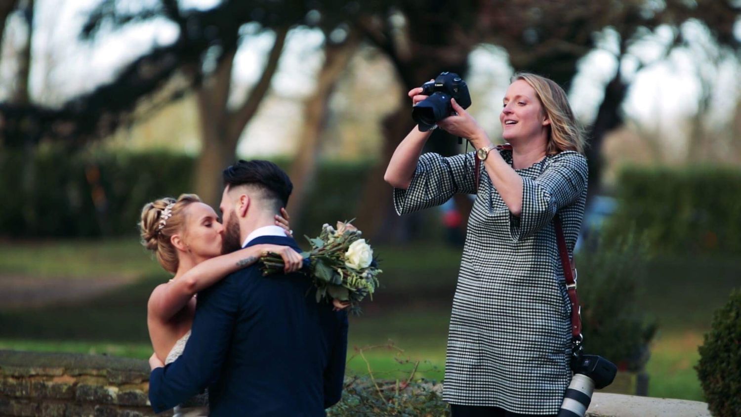 Wedding photographer capturing a couple kissing outdoors, bride holding a bouquet, at a garden venue.