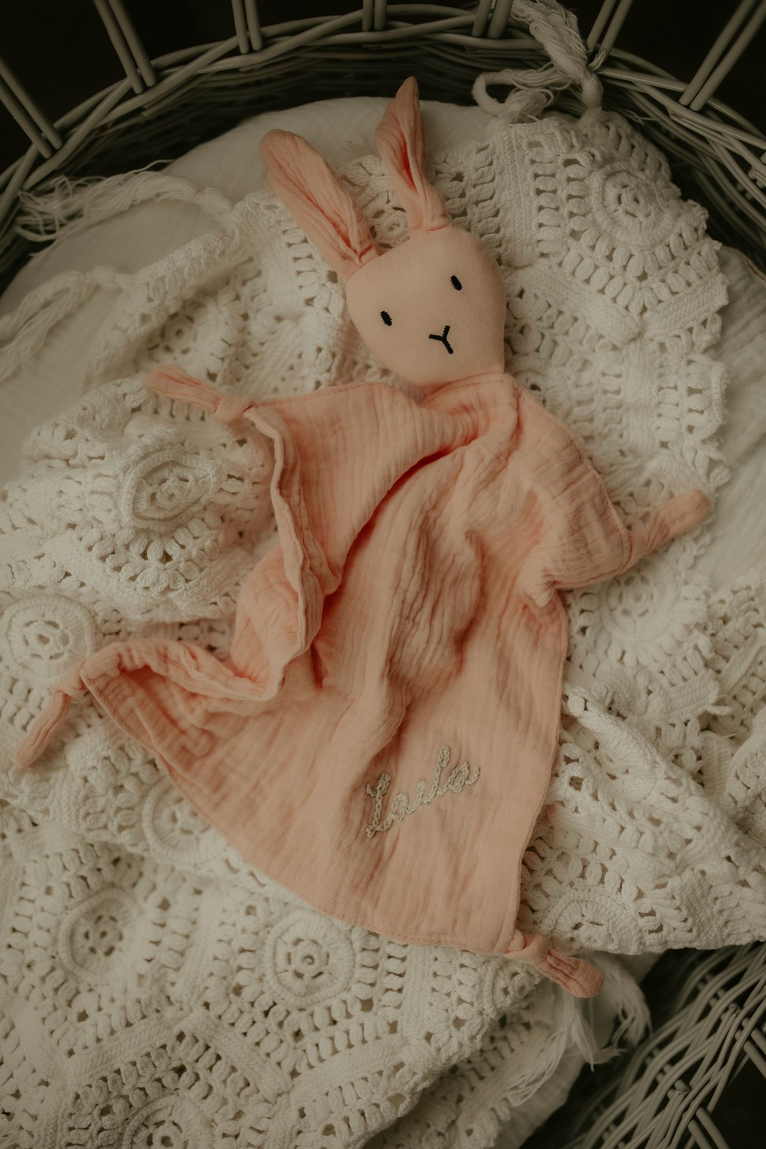 Pink bunny lovey security blanket toy resting in a wicker basket lined with white lace fabric.