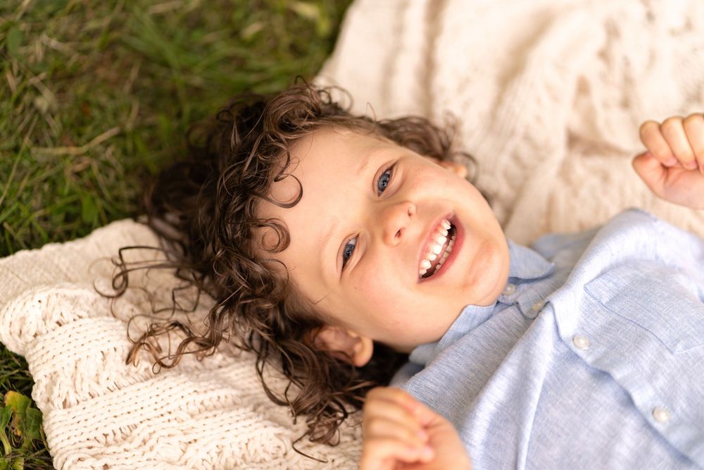 Smiling curly-haired child lying on a knit blanket in green grass, wearing a light blue shirt.