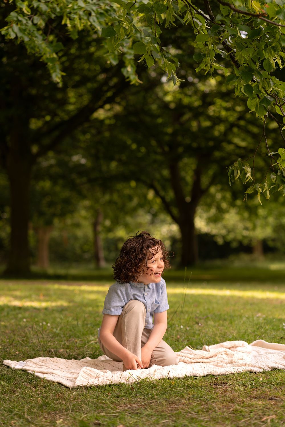 A curly-haired child sits on a blanket in a sunny park, surrounded by lush green trees.