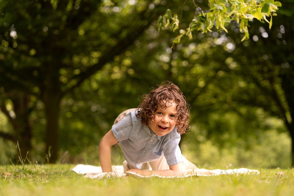A happy curly-haired child lying on a blanket in a sunny green park, smiling joyfully at the camera.
