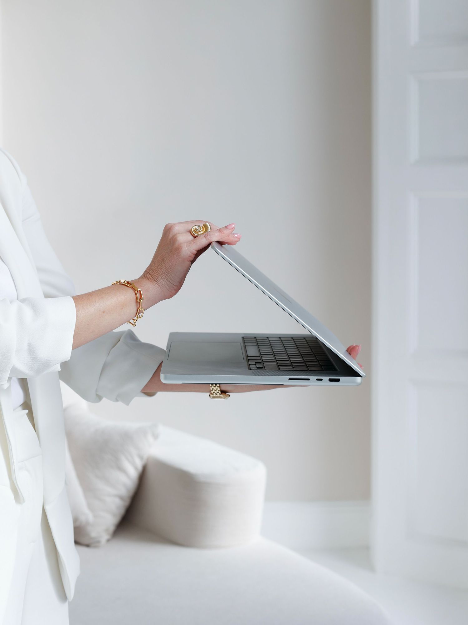 A woman in a white outfit holds an open silver laptop with gold jewelry on her wrist in a bright minimalist room.
