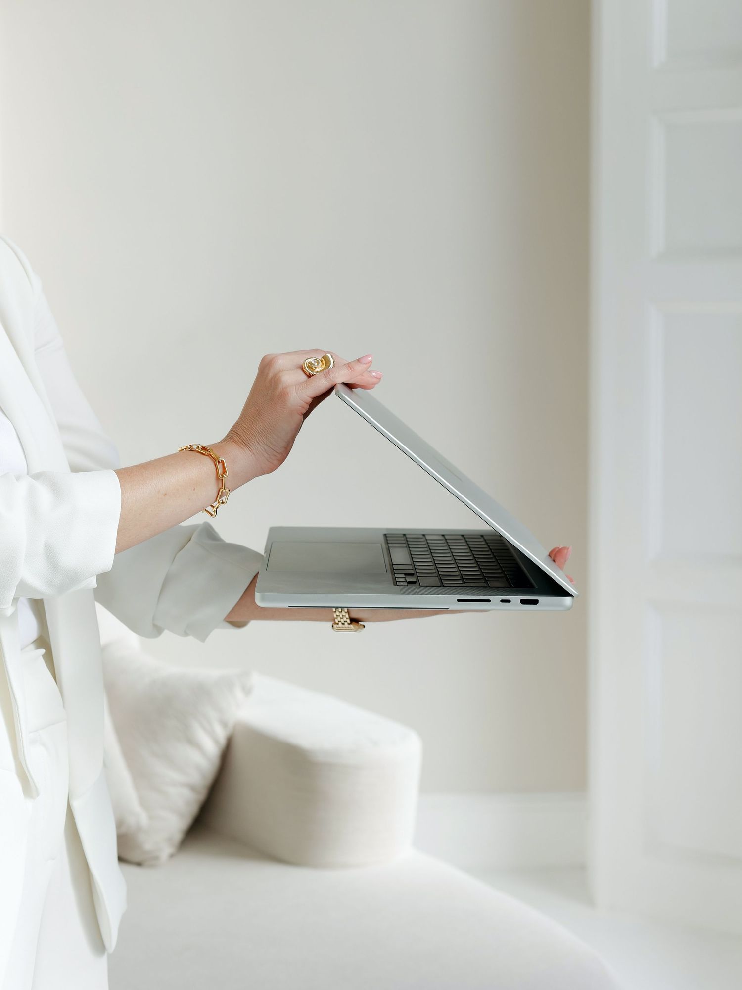 A woman in a white outfit holds an open silver laptop with gold jewelry on her wrist in a bright minimalist room.