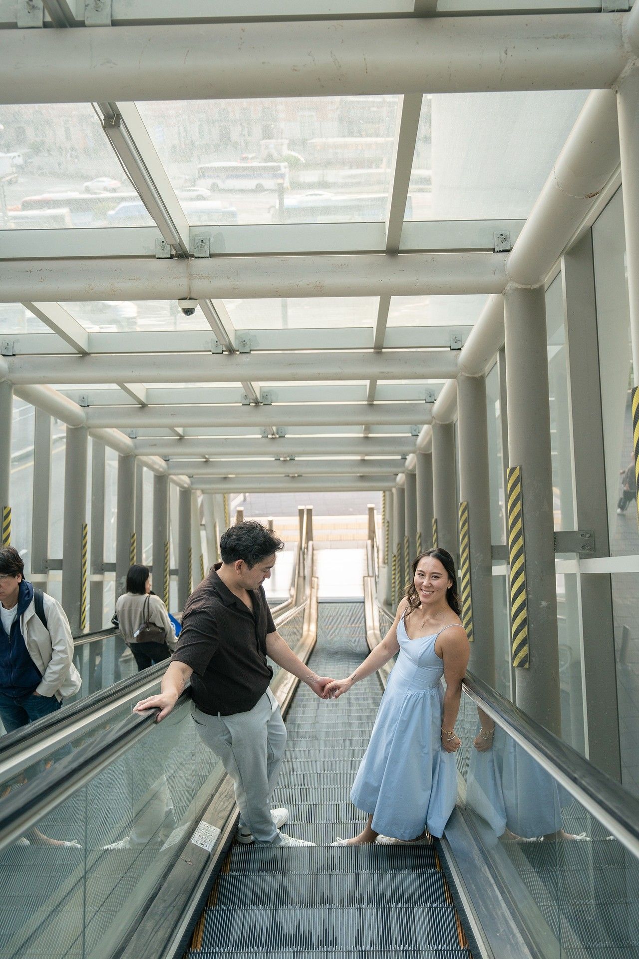 Couple holding hands on an escalator in a modern glass corridor, woman in blue dress, man in black shirt.