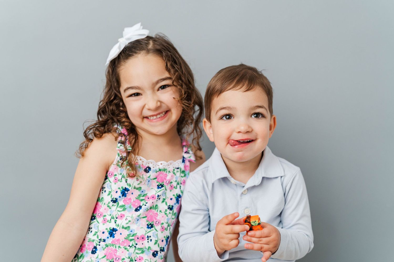 Two smiling young children posing together, a girl in a floral dress and a boy in a blue shirt, against a gray background.