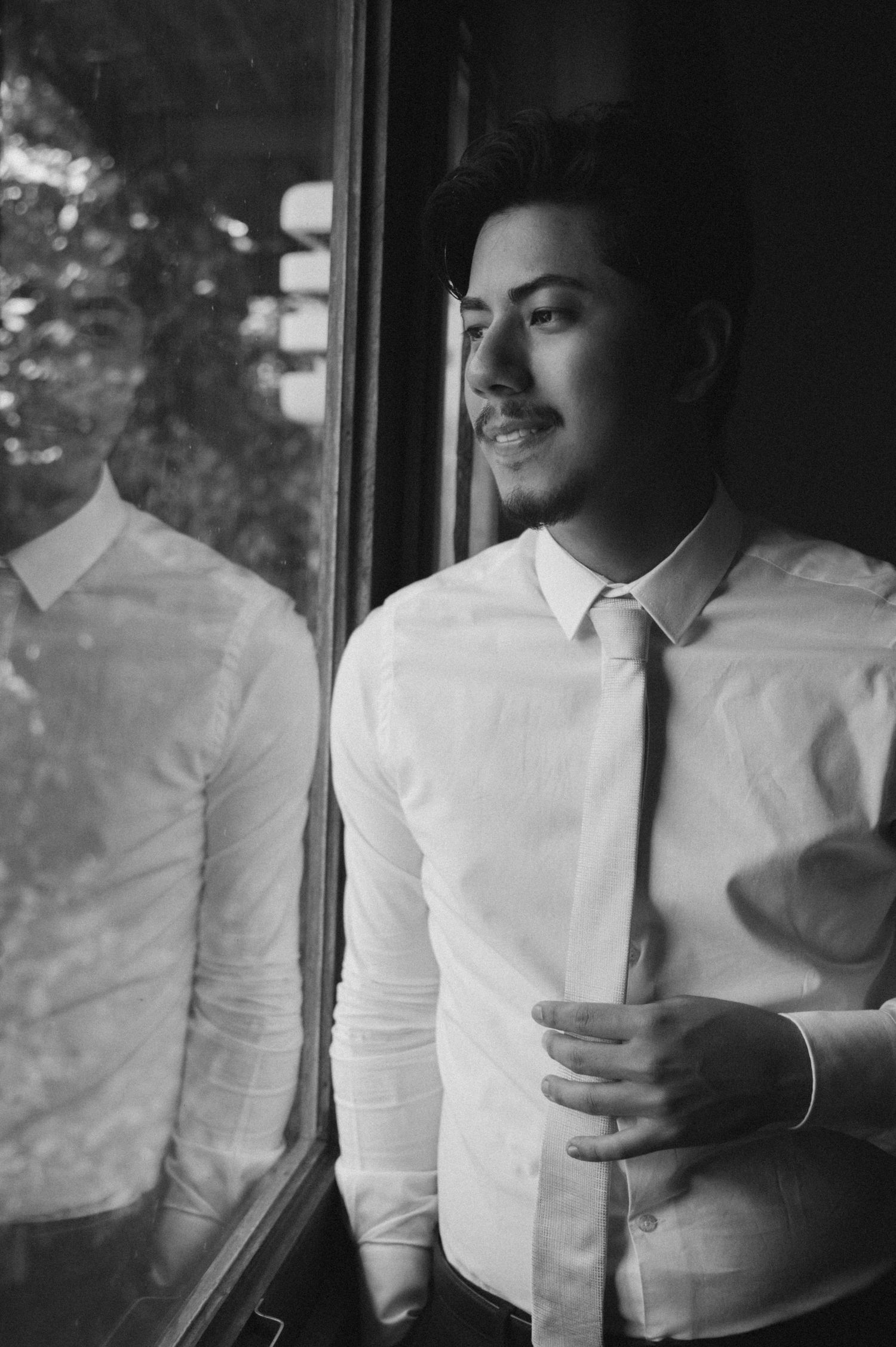 Black and white portrait of a young man in white shirt and tie gazing thoughtfully out a window with his reflection visible.