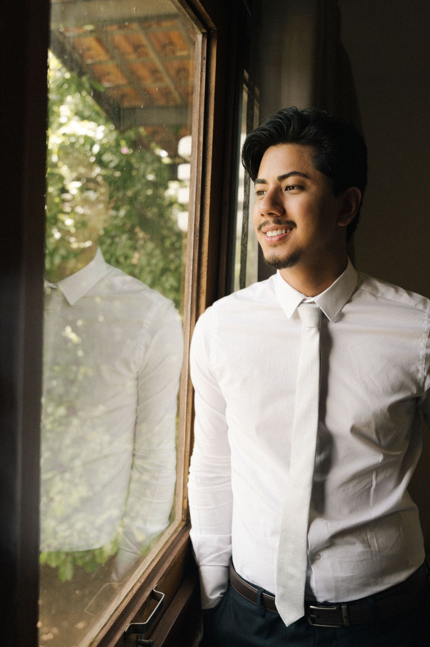 Smiling young man in white dress shirt and tie standing by a sunlit window with green trees reflected in the glass.