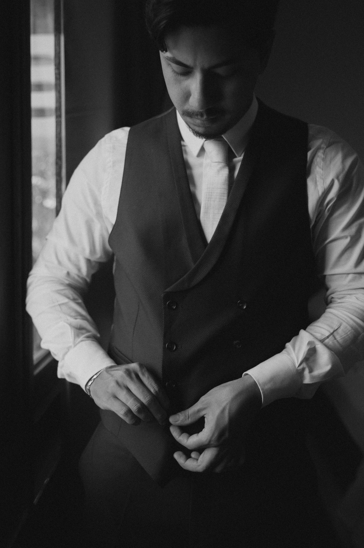 Groom in white dress shirt and dark vest buttoning his jacket in moody black and white wedding preparation photo.