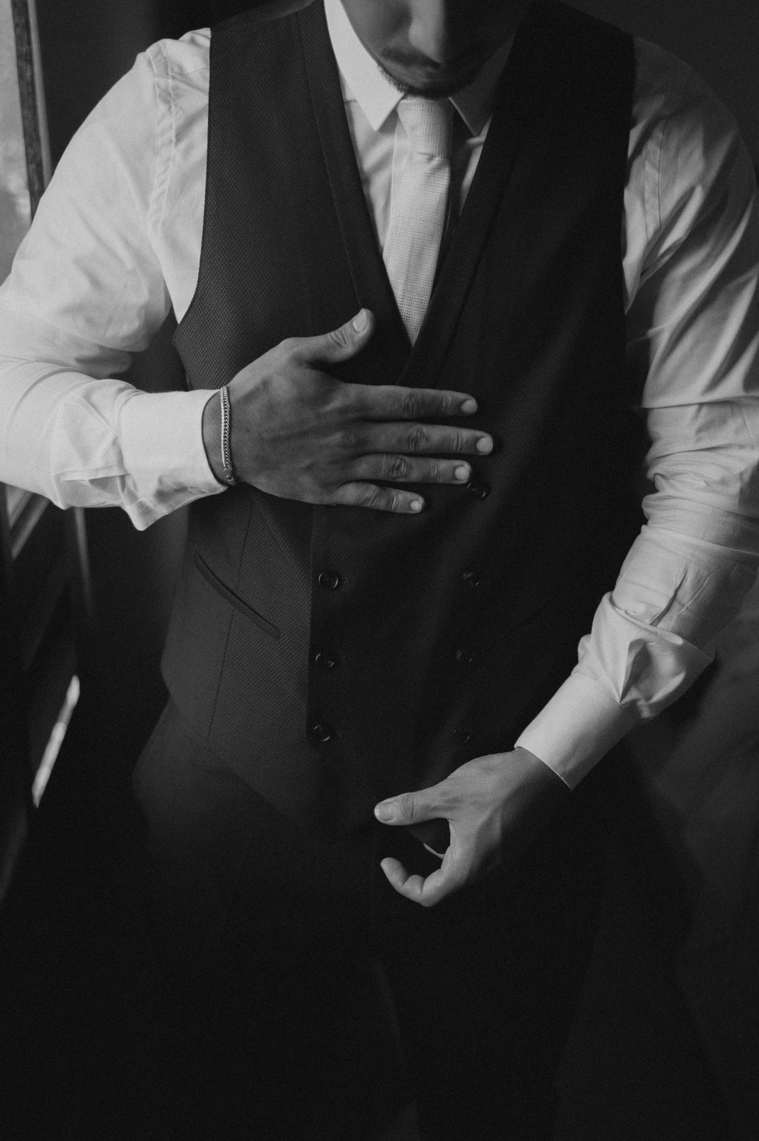 Black and white photo of a groom in a white shirt, tie, and vest, adjusting his suit before the wedding ceremony.