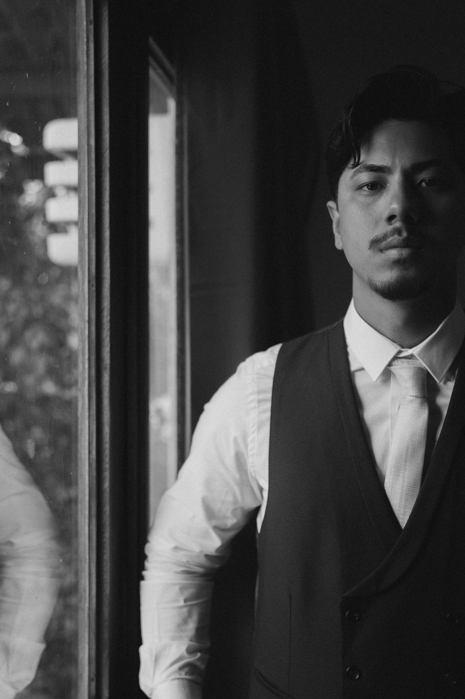 Black and white portrait of a young man in a vest and tie standing by a reflective window.