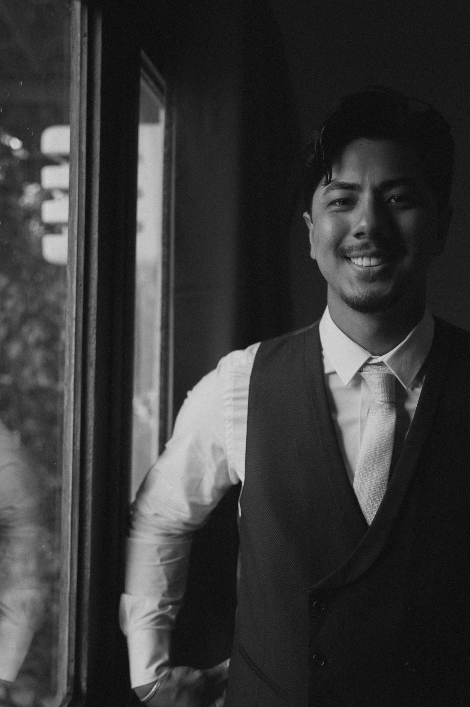 Smiling man in a vest and tie standing by a window in a black and white portrait photo.