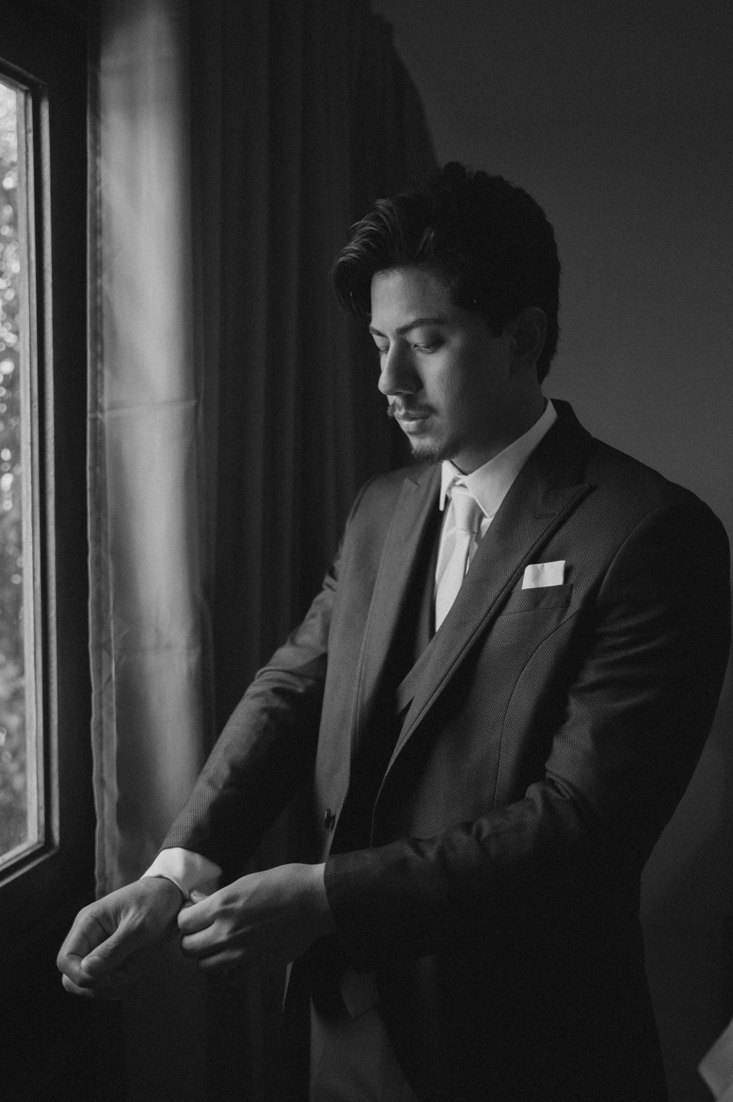 Black and white photo of a well-dressed man in a suit adjusting his cufflinks by a window.