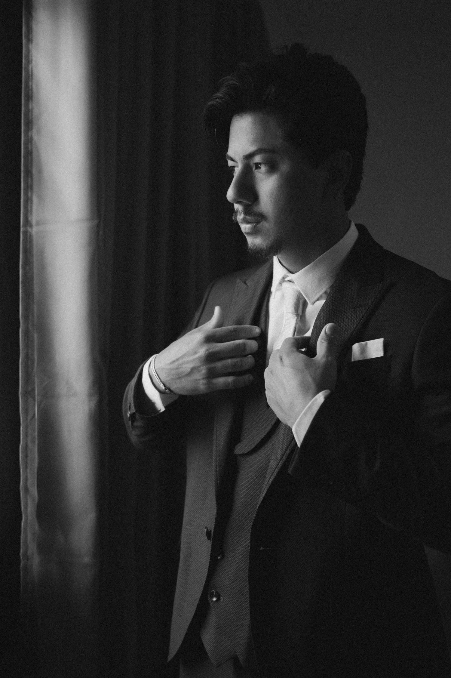 Black and white portrait of a well-dressed groom in a suit adjusting his tie near a window.