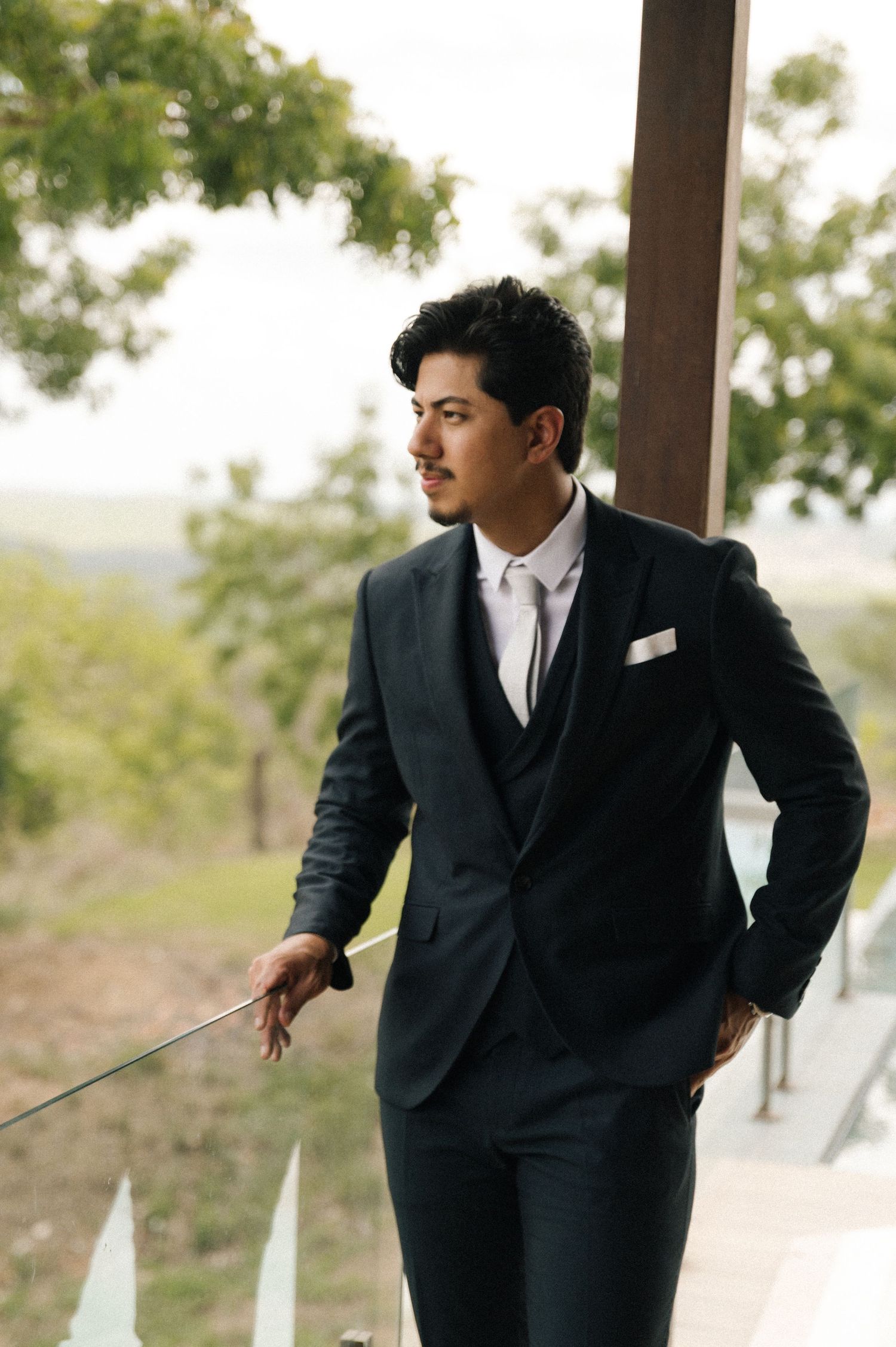 Stylish man in dark three-piece suit with white tie and pocket square standing on outdoor balcony.