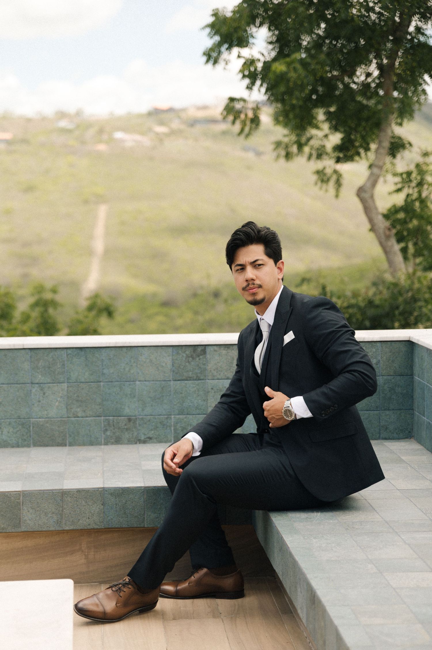 Man in black suit and brown shoes sitting on tiled steps with green hillside background.