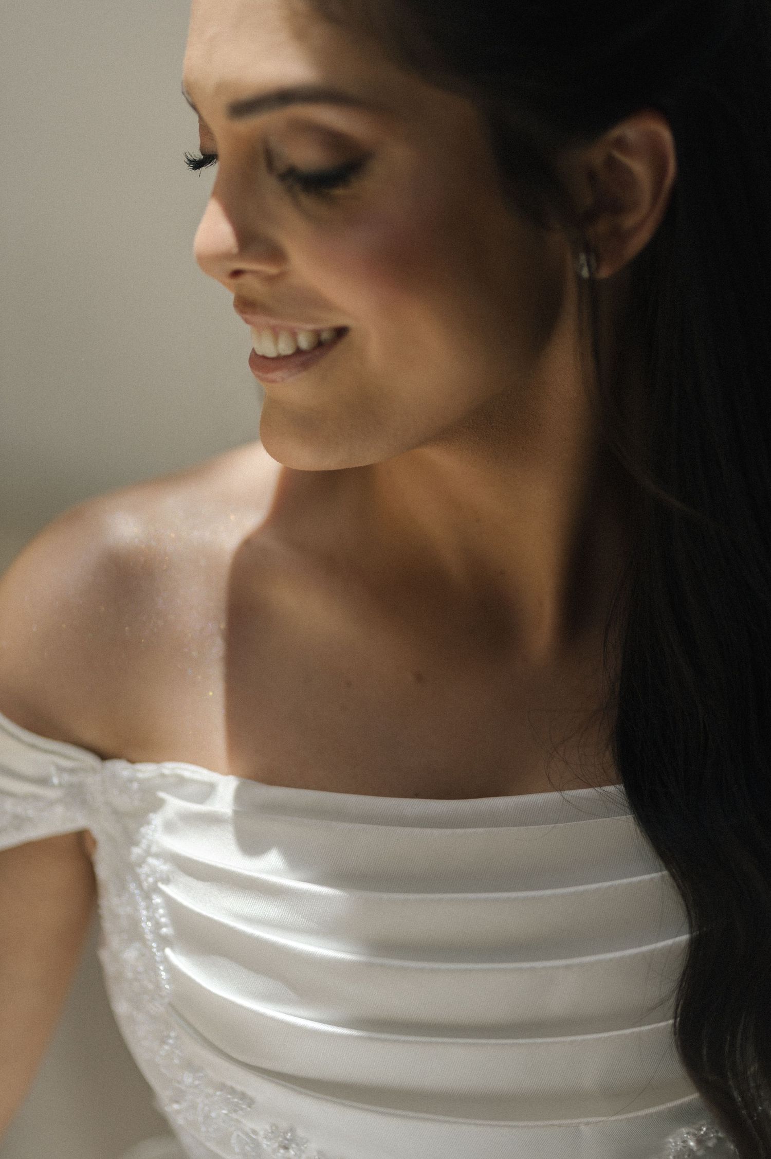 Smiling bride in an off-shoulder white wedding gown with elegant makeup and dark hair, captured in soft natural light.