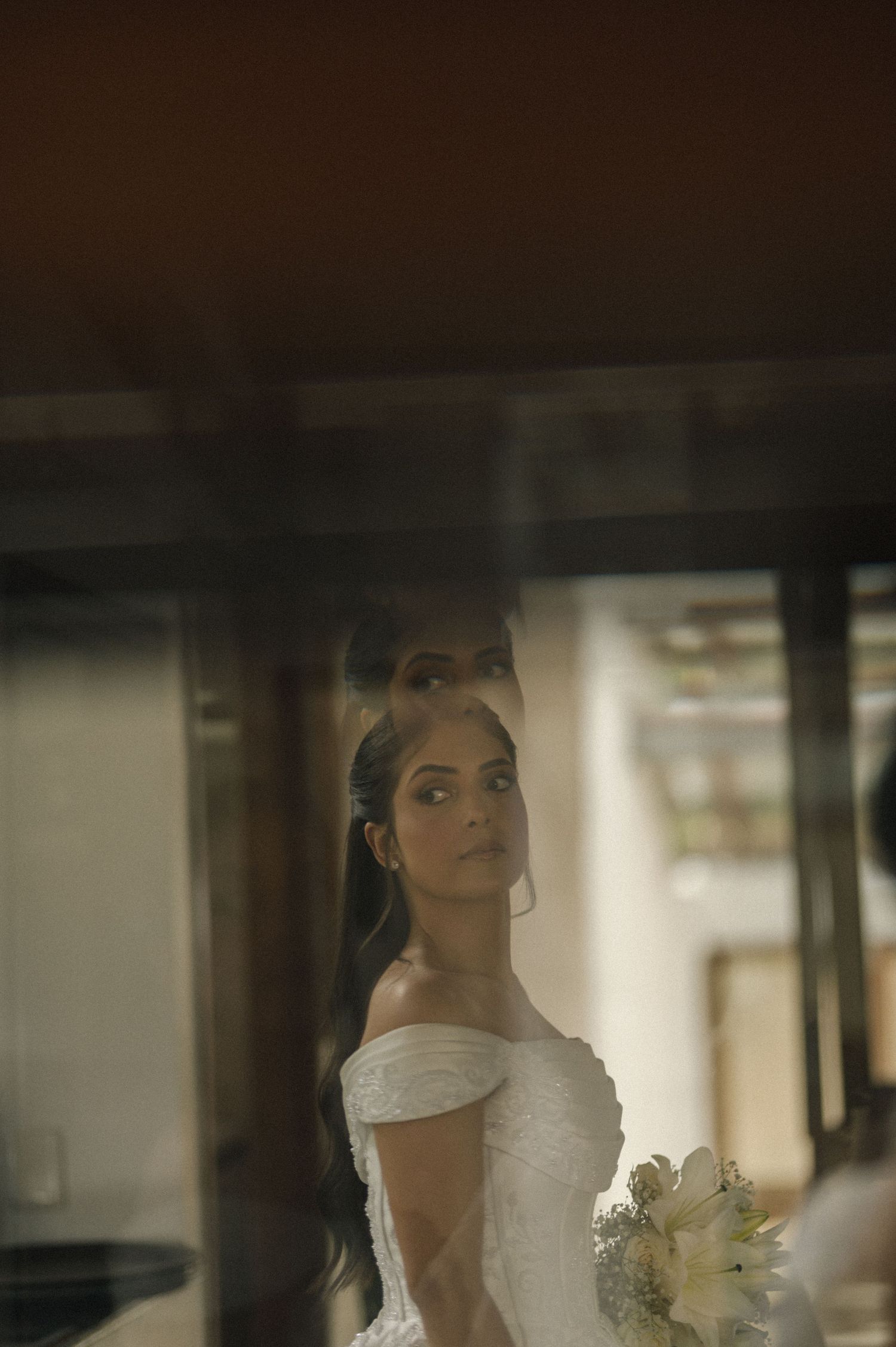 A bride in an off-shoulder white gown holds a bouquet, her reflection visible in a window.