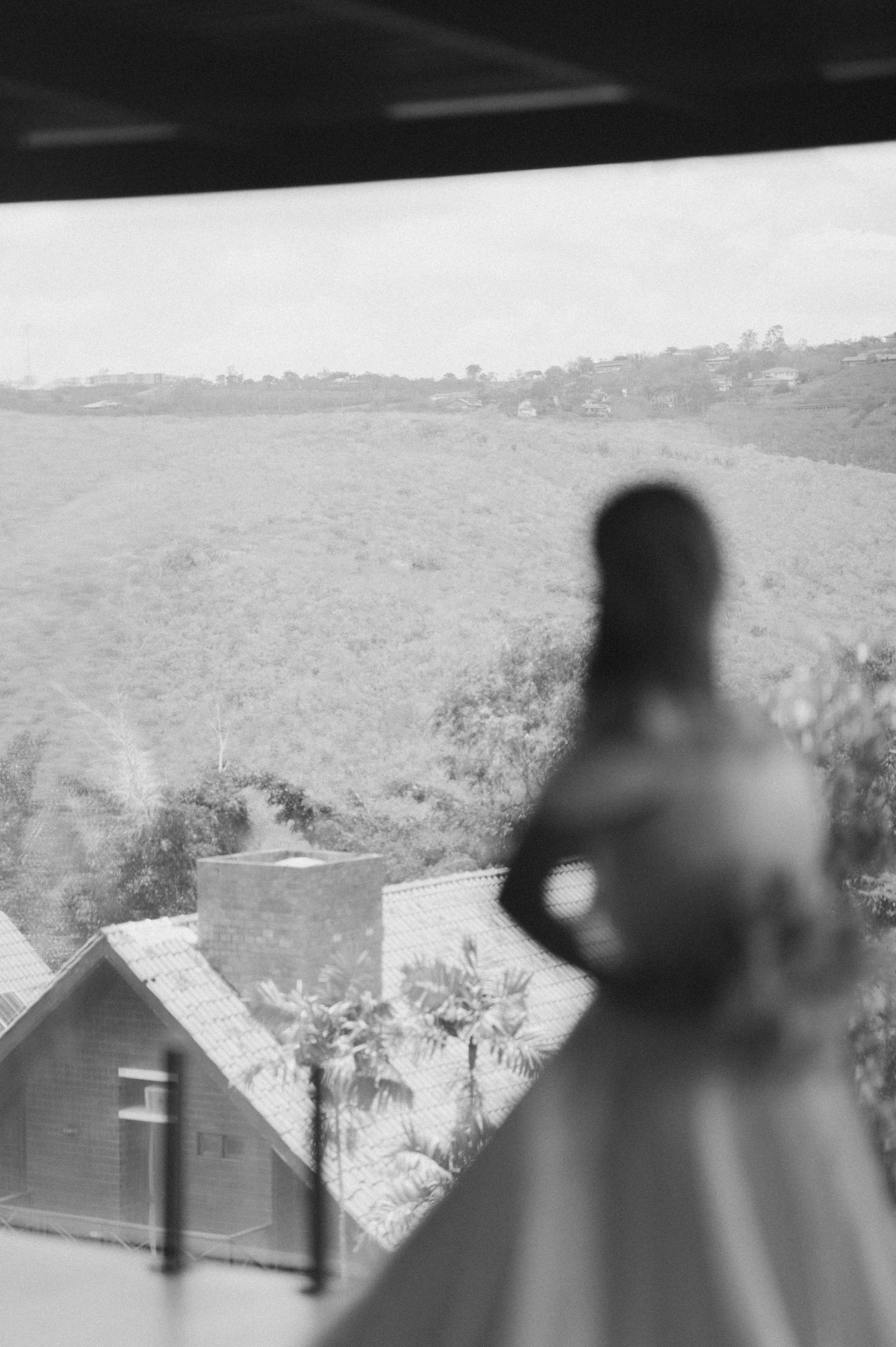 Black and white photo of a blurred bride holding bouquet, gazing out window at rolling green hills and rustic buildings.