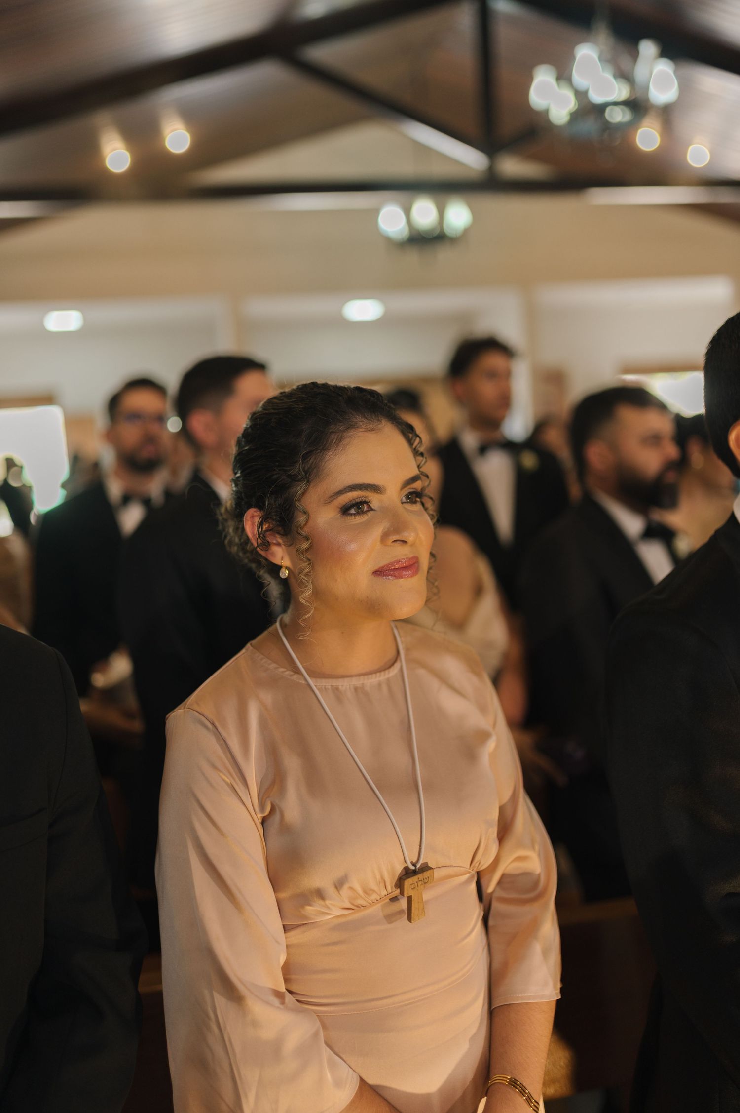 Elegant woman in blush dress with cross necklace watching a wedding ceremony among guests.