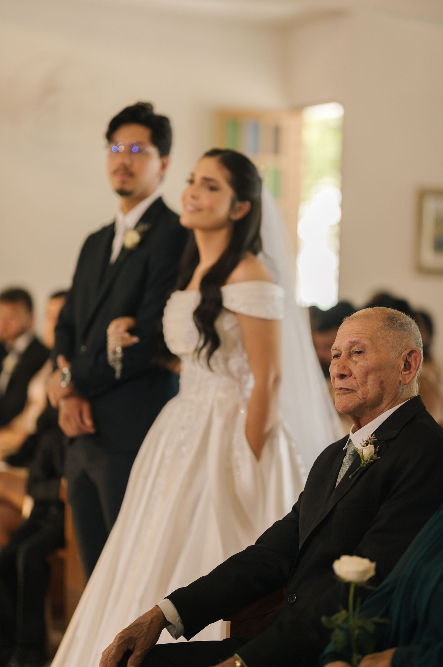 Bride and groom stand at the altar during a church wedding ceremony, with an elderly guest seated in the foreground.