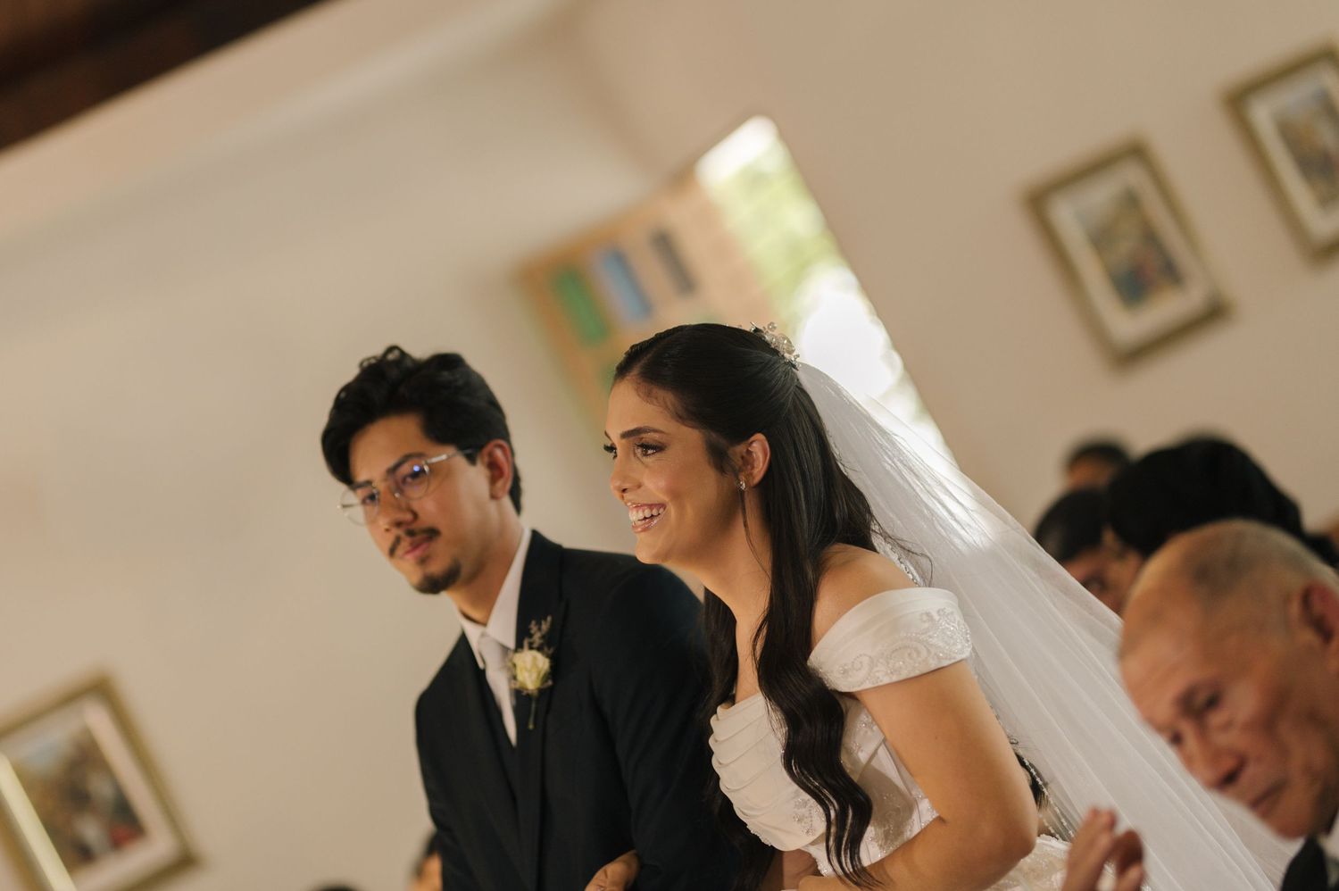 Bride in white veil smiles beside groom in black suit during wedding ceremony in a church.