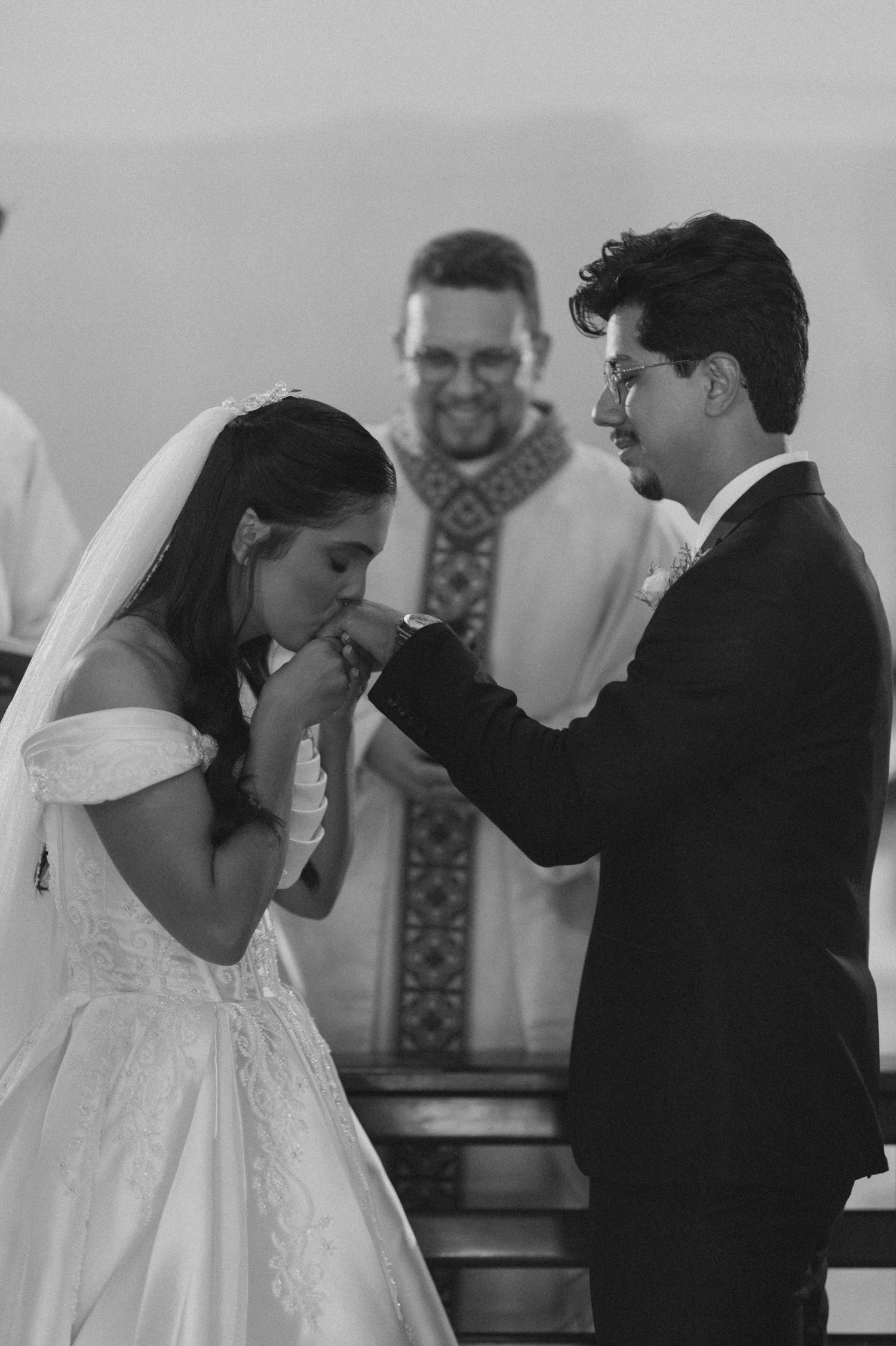 Black and white wedding photo of bride kissing grooms hand at altar with smiling officiant in background.