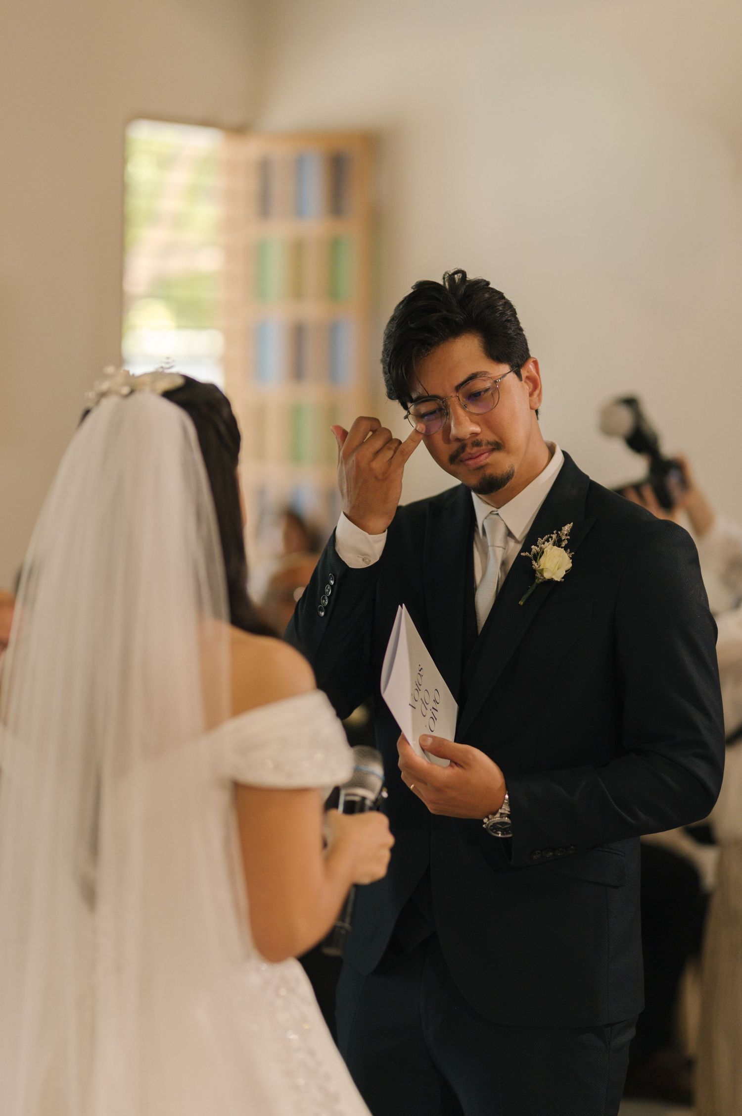Emotional groom wiping tears during wedding ceremony while bride reads vows in a church.