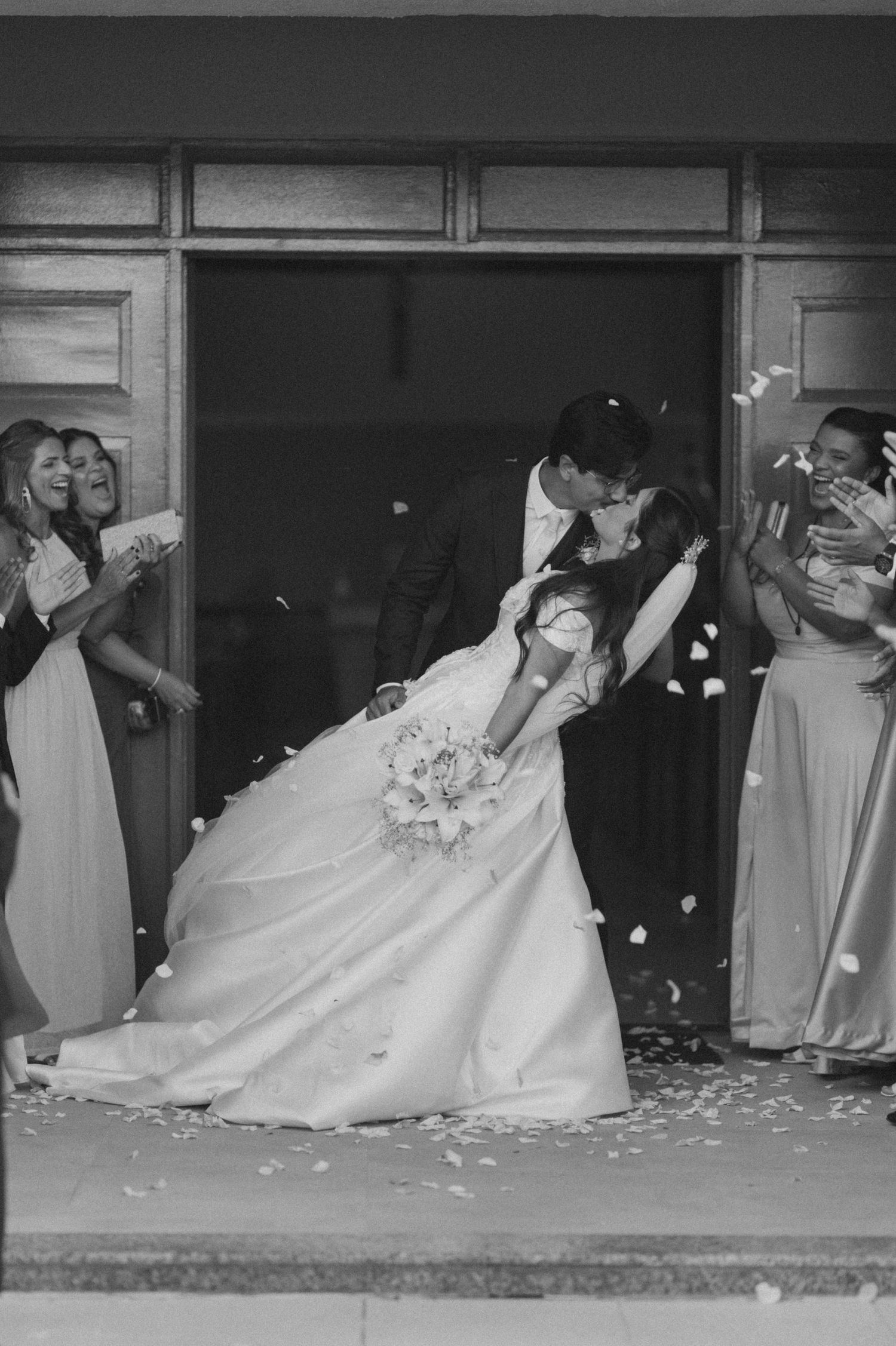 Black and white photo of a bride and groom sharing a romantic dip kiss outside a church, surrounded by bridesmaids tossing petals.