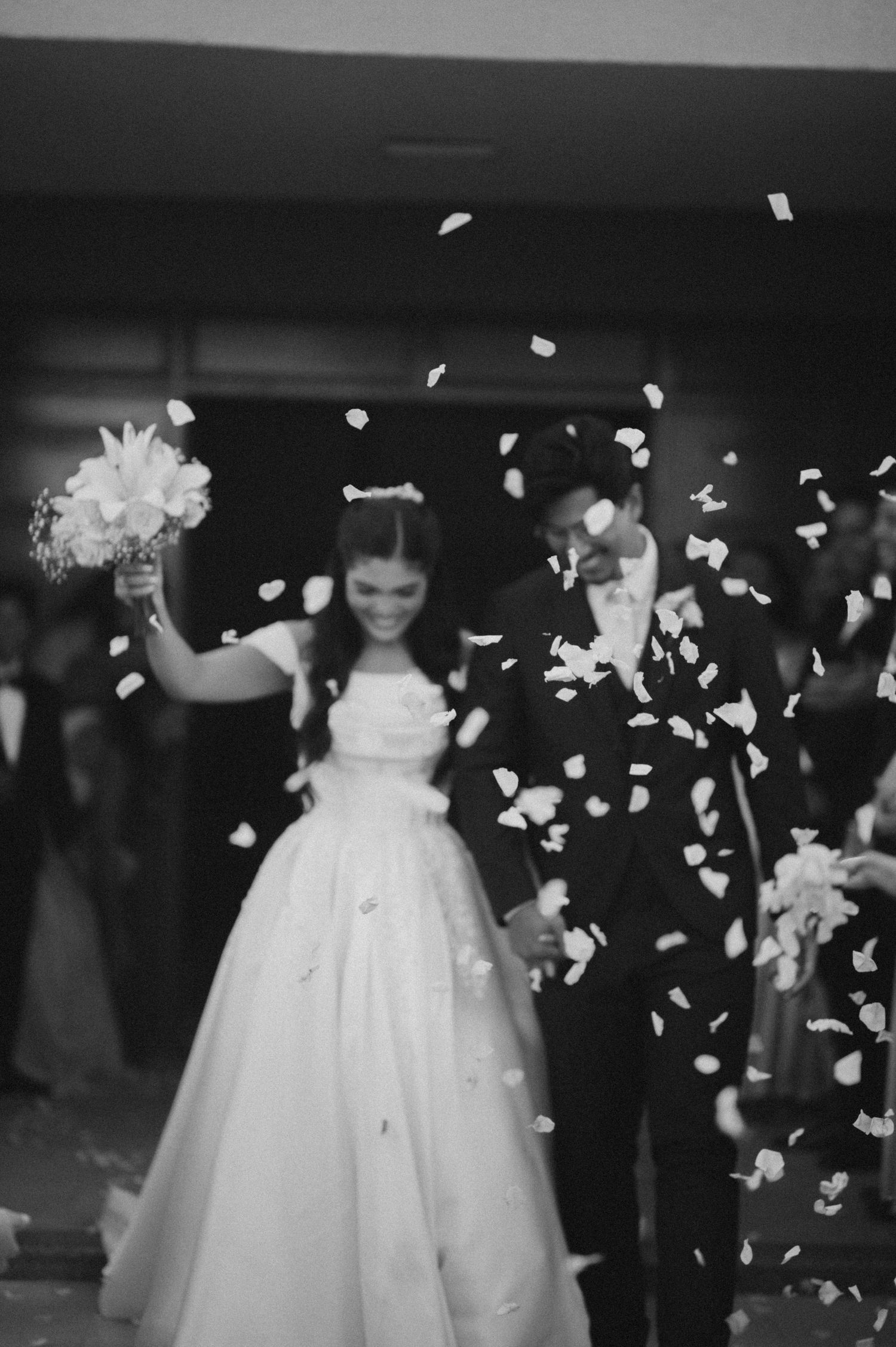 Black and white photo of a bride and groom walking through a shower of flower petals after their wedding ceremony.