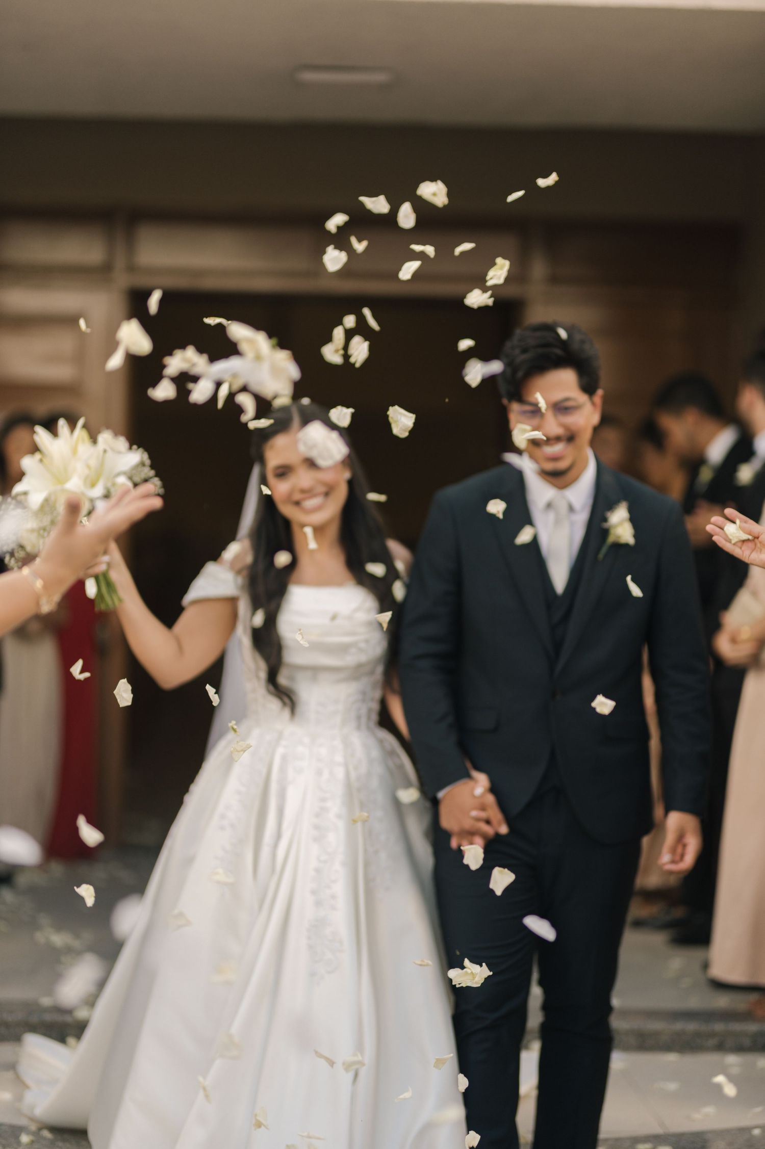 Newlywed couple walking out smiling as white flower petals are tossed in celebration after their wedding ceremony.