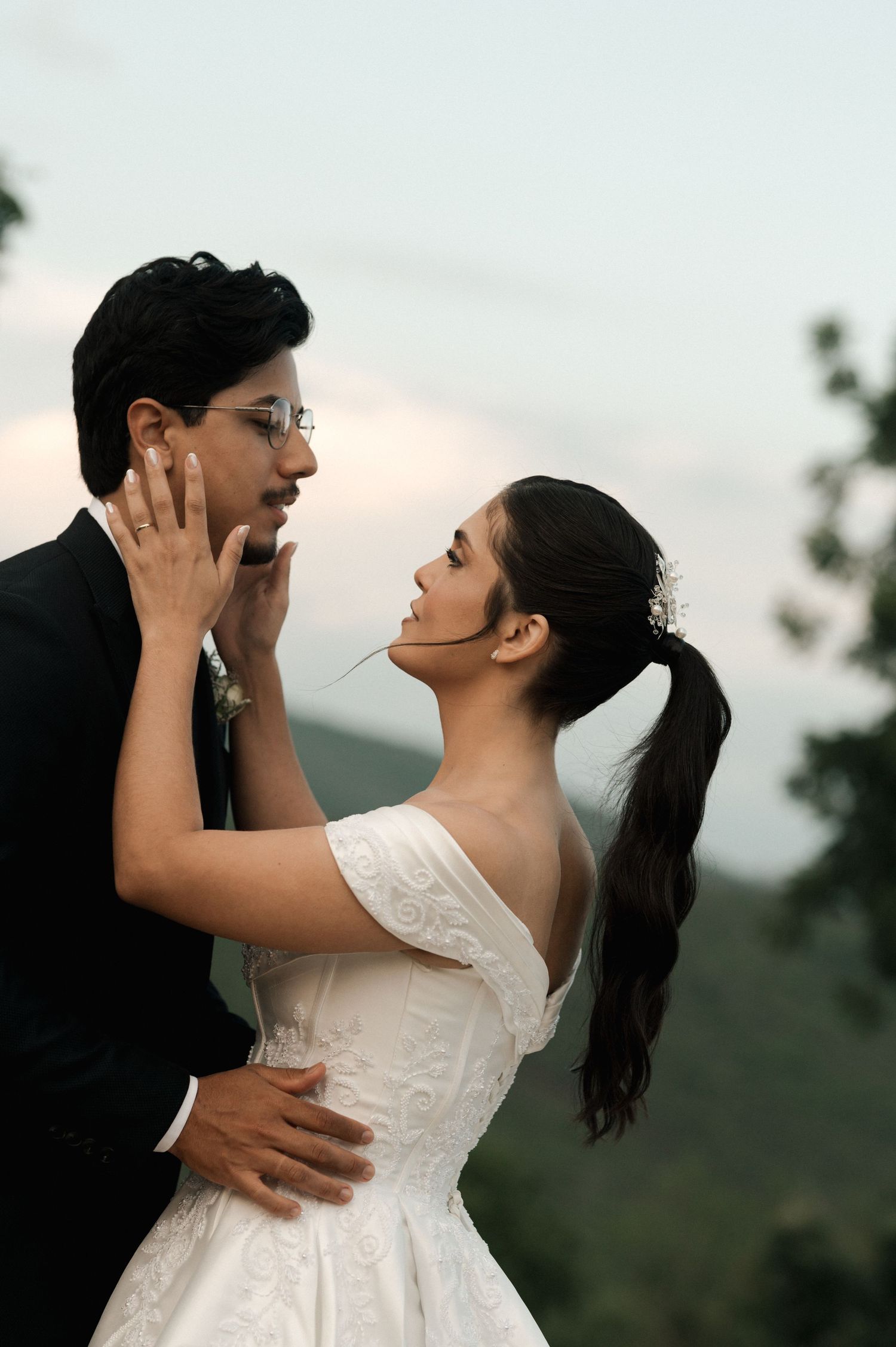 A bride in a white gown tenderly holds a groom's face outdoors, sharing a romantic wedding moment.