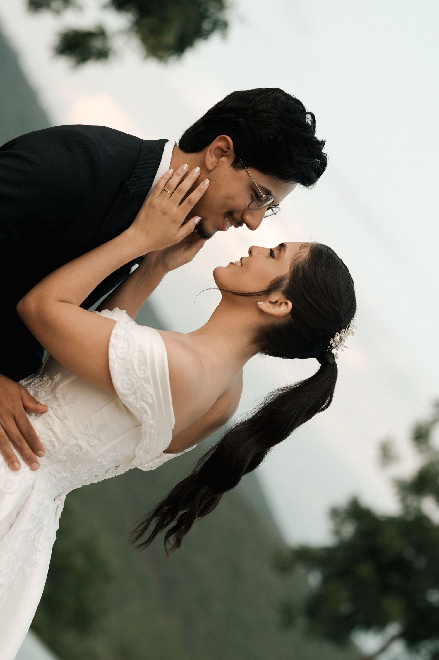 A couple in formal attire share a romantic moment outdoors, with the man gently holding the woman's face.