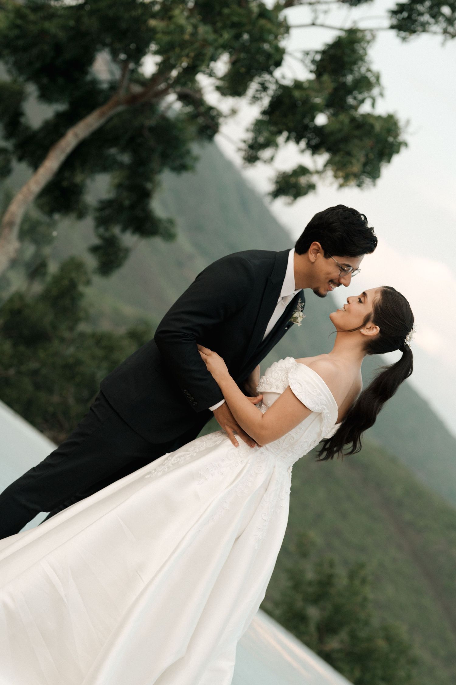 Bride and groom sharing a romantic dip kiss outdoors with lush green mountains in the background.