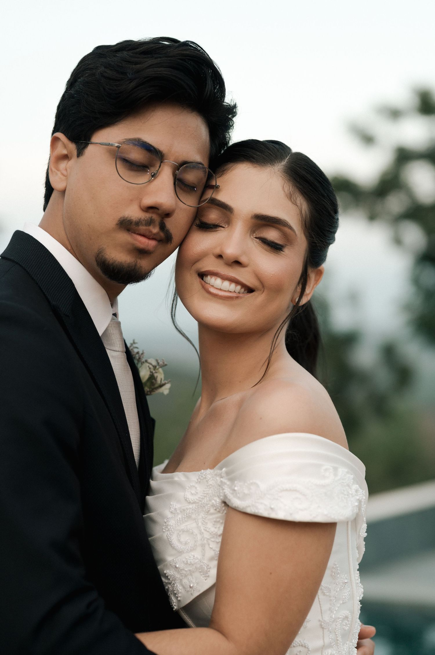 Smiling bride in off-shoulder gown embraces groom in glasses during outdoor wedding portrait.