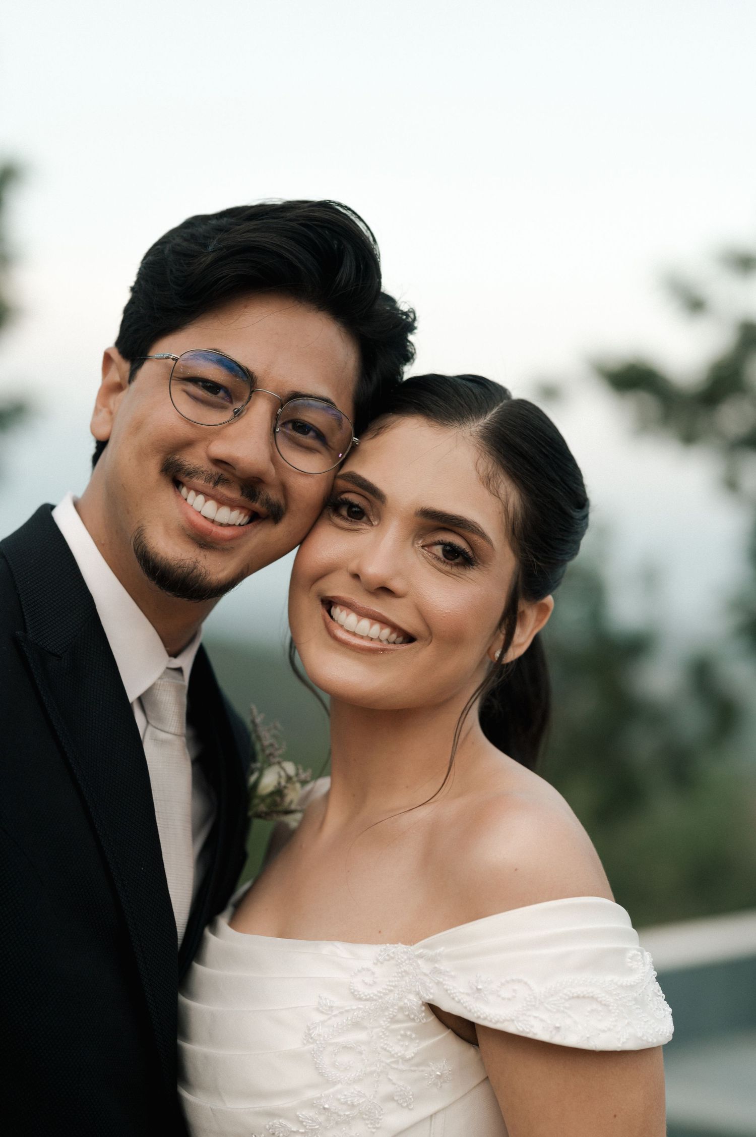 A smiling couple in wedding attire, groom in black suit with glasses, bride in white off-shoulder dress, posing outdoors.
