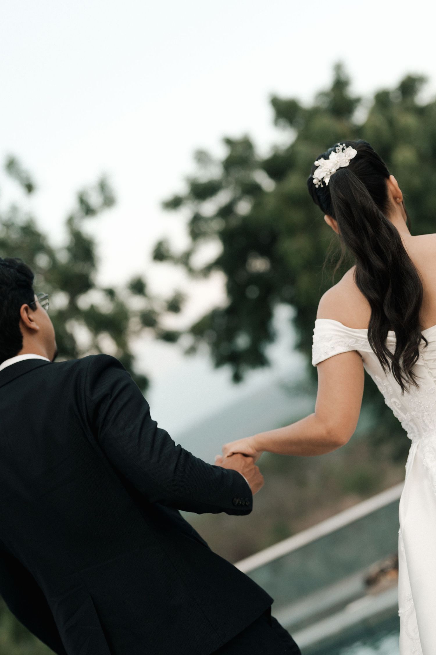 A bride and groom hold hands outdoors, she wears an off-shoulder white gown with floral hair accessory.